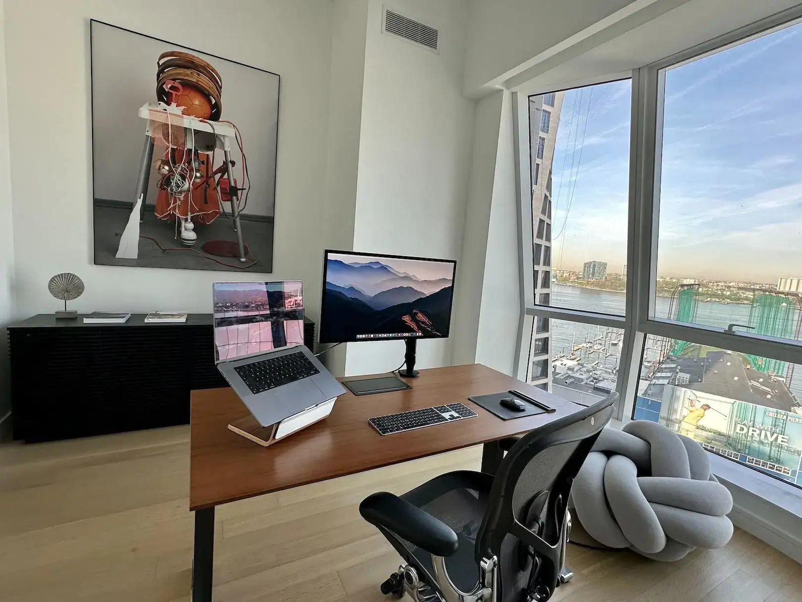 Laptop with dual screens and ergonomic chair in modern home office, city skyline view.